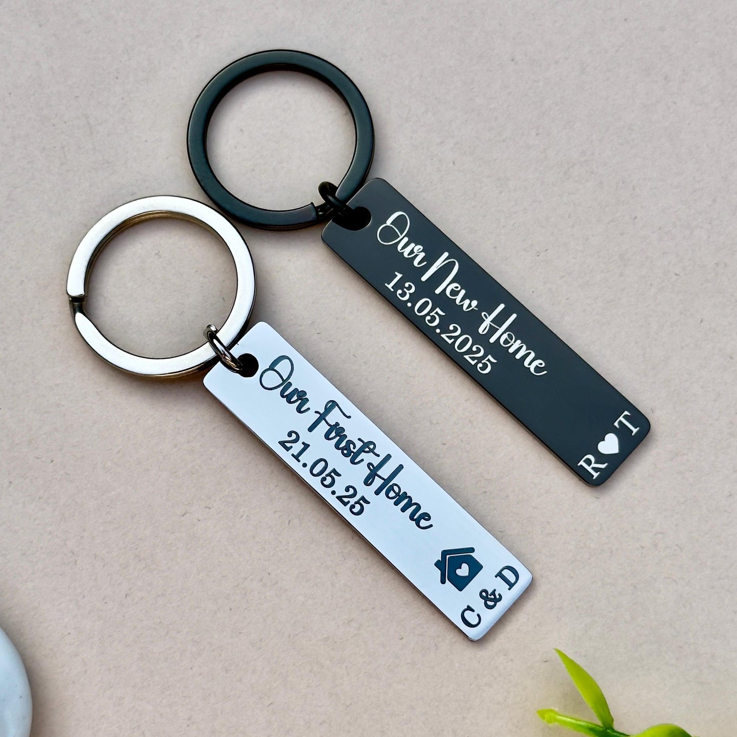 et of two personalized engraved stainless steel keychains, perfect for celebrating home milestones. The black keychain features 'Our New Home 13.05.2025' and initials 'R.T.' with a heart symbol, ideal for couples. The silver keychain is engraved with 'Our First Home 21.05.25' and initials 'C.D.' with a house symbol, making it a great gift for first-time homeowners. Both keychains are made from durable stainless steel, attached to sturdy metal rings, and feature a