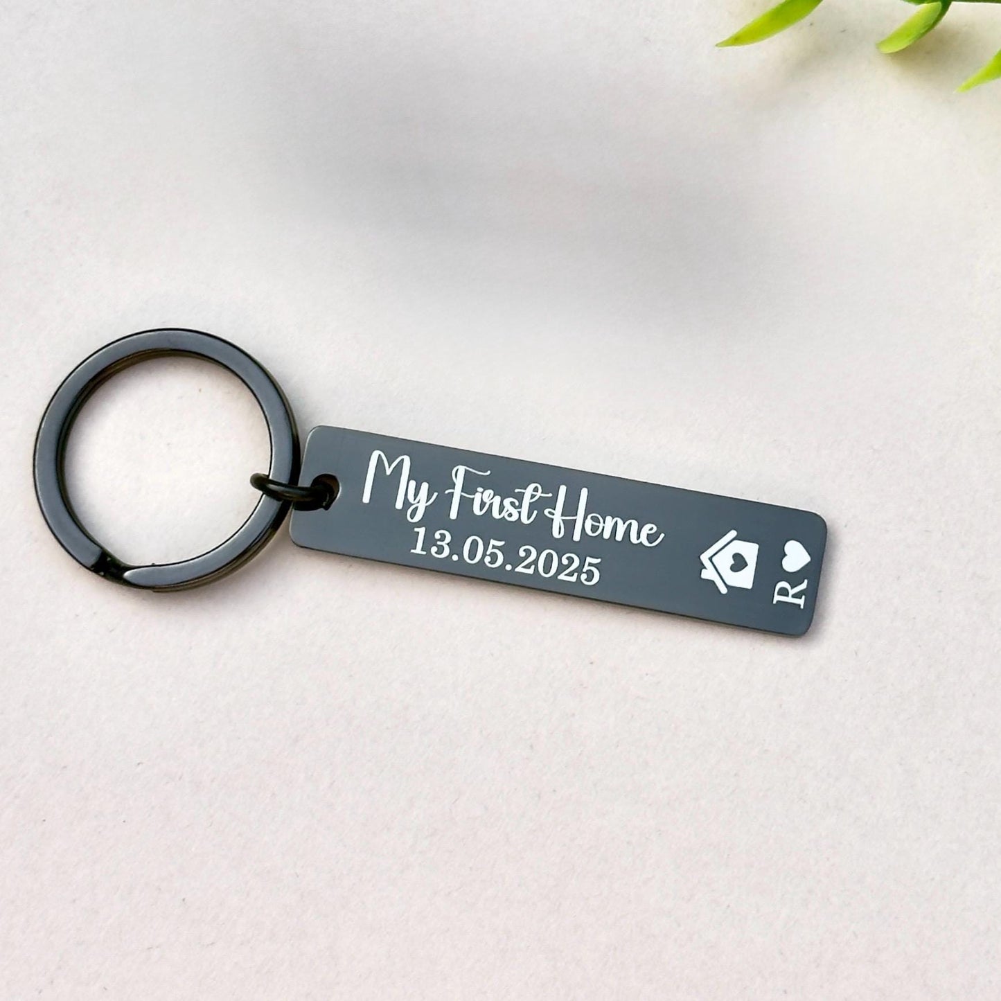 Personalized black stainless steel keychain engraved with 'My First Home 13.05.2025' and the initials 'R,' along with a house symbol and a heart symbol. This sleek and modern keychain makes an ideal gift for individuals or couples celebrating their first home purchase. Crafted from high-quality stainless steel, it features a polished finish and is attached to a sturdy metal ring, perfect for keys or as a keepsake