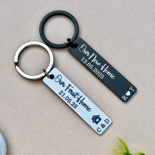et of two personalized engraved stainless steel keychains, perfect for celebrating home milestones. The black keychain features &#39;Our New Home 13.05.2025&#39; and initials &#39;R.T.&#39; with a heart symbol, ideal for couples. The silver keychain is engraved with &#39;Our First Home 21.05.25&#39; and initials &#39;C.D.&#39; with a house symbol, making it a great gift for first-time homeowners. Both keychains are made from durable stainless steel, attached to sturdy metal rings, and feature a