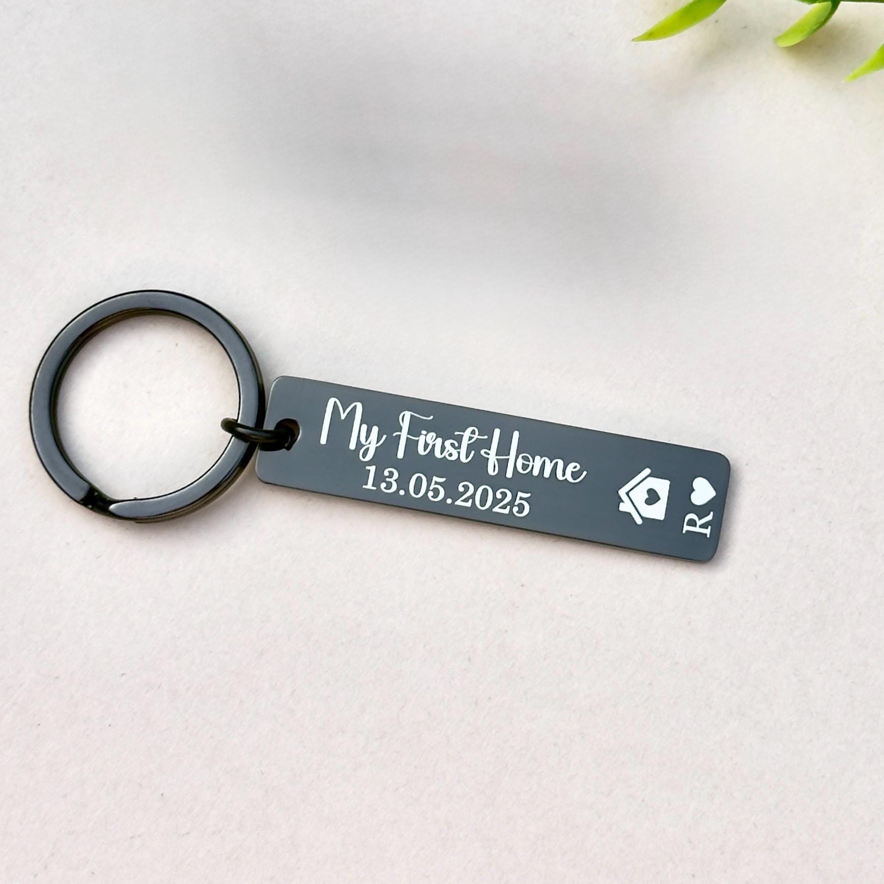 Personalized black stainless steel keychain engraved with &#39;My First Home 13.05.2025&#39; and the initials &#39;R,&#39; along with a house symbol and a heart symbol. This sleek and modern keychain makes an ideal gift for individuals or couples celebrating their first home purchase. Crafted from high-quality stainless steel, it features a polished finish and is attached to a sturdy metal ring, perfect for keys or as a keepsake